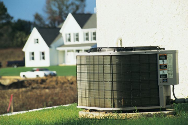 An outdoor air conditioning unit sits on a concrete pad next to a white house, with another white house and a car visible in the background across a grassy lawn.