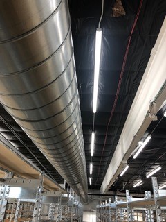 A large metal air duct runs along the ceiling of an industrial space with fluorescent lights and metal shelving units below. The ceiling is partially open, revealing pipes and wiring.