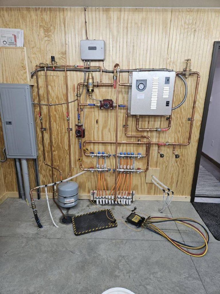 A complex arrangement of copper pipes, valves, gauges, and control panels mounted on a wooden wall in a utility room, with hoses and a small metal tank on the concrete floor below.