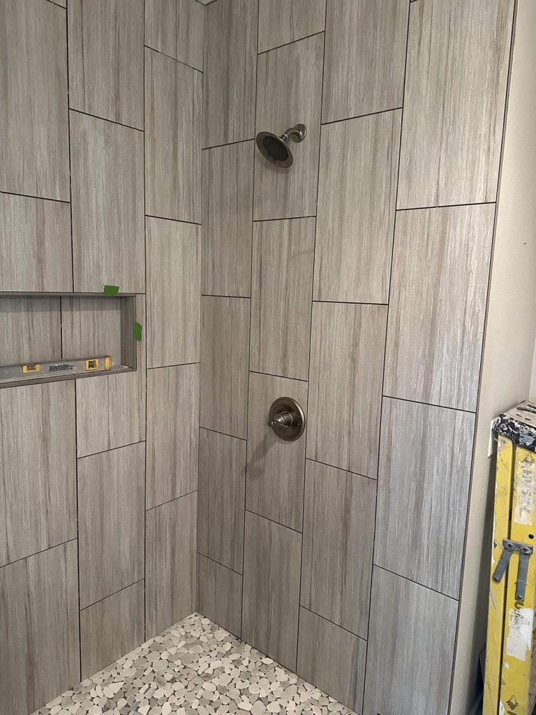 1e1fe74e-f7da-4e9d-a415-b1cdfdac3895 A shower with gray rectangular wall tiles, a built-in shelf holding a level tool, two metal fixtures, and a floor with small, irregular white and gray tiles. Part of a yellow construction tool is visible on the right.