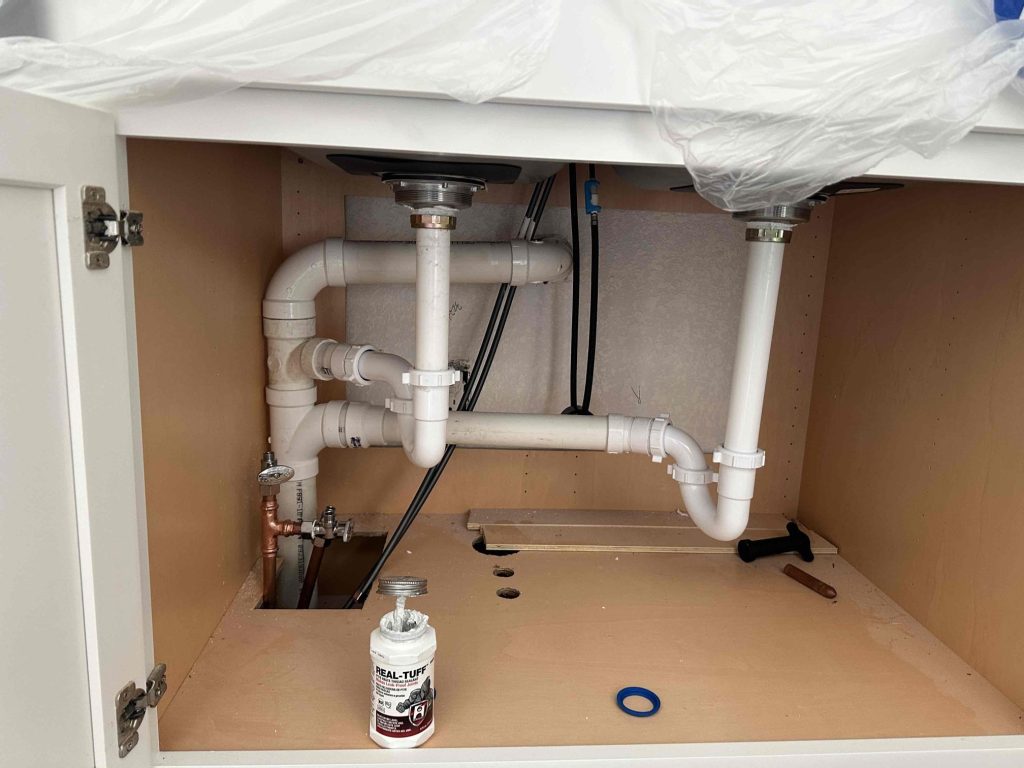 Plumbing pipes under a sink inside a cabinet, with white PVC pipes, fittings, a bottle of pipe cement, a blue washer, and plumbing tools placed on the wooden cabinet floor.