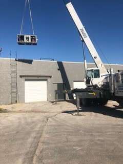 A crane lifts a large HVAC unit onto the roof of a gray industrial building with white garage doors on a clear, sunny day.