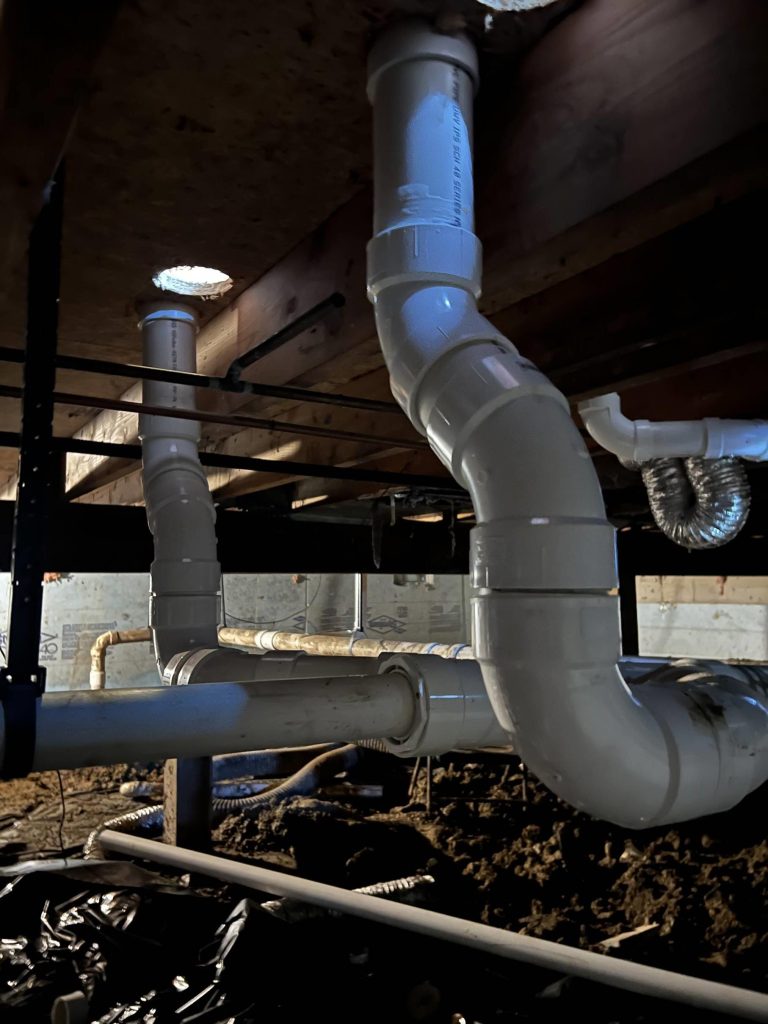 Large white PVC pipes and fittings run along the ceiling and floor of a dimly lit crawlspace, with exposed soil and plastic sheeting on the ground. The pipes are connected with joints and elbows, part of a plumbing system.