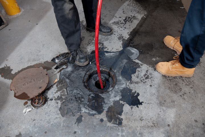 Two people stand near an open manhole with a red hose inserted inside; oil stains and a removed manhole cover are visible on the concrete ground.