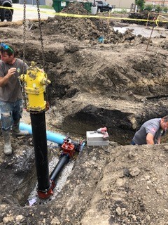 Two construction workers install a yellow fire hydrant attached to a black pipe in a large dirt excavation site, with tools and equipment nearby.