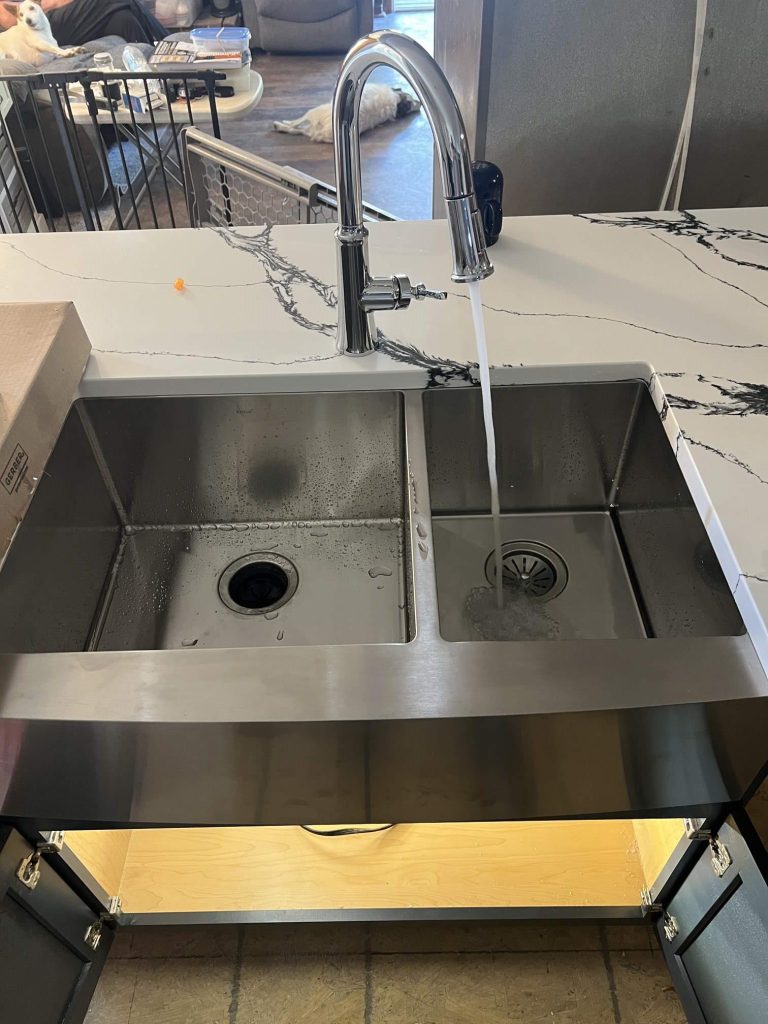 6642c7f7-a578-40b3-948c-02ee8f7e5e3f A modern double-basin stainless steel kitchen sink with a tall faucet running water into the right basin, set in a white marble countertop with black veins. The under-sink cabinet is open, revealing a wooden base.