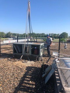 A worker stands on a gravel-covered rooftop near a large HVAC unit being hoisted by a crane, under a clear blue sky. Tools and equipment are scattered nearby.