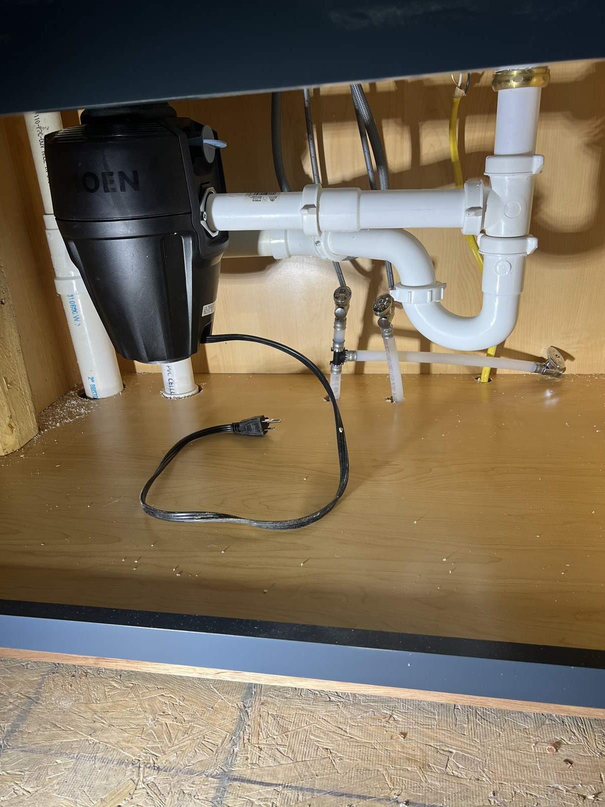 A garbage disposal unit under a kitchen sink with white PVC pipes, various water lines, and a black electrical cord unplugged, lying on the wooden cabinet floor.