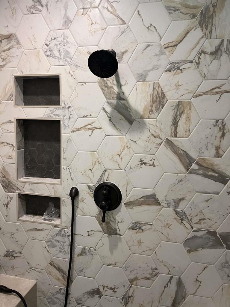 93f7d546-7ec1-47bf-8ba8-b143bb3c07ae A shower wall with large marble hexagon tiles, a black showerhead, a black control handle, a handheld shower attachment, and two built-in wall niches with smaller hexagon tiles inside.