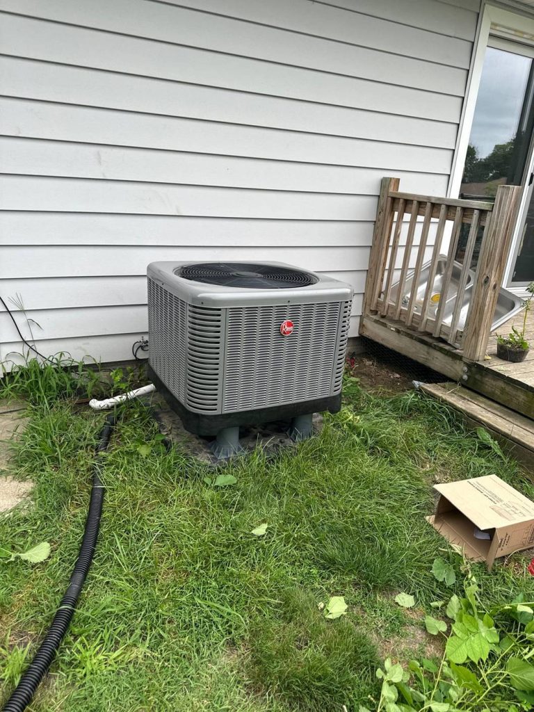 A gray outdoor air conditioning unit sits on a concrete pad next to a white-sided house with a small wooden porch. The surrounding grass is slightly overgrown, and an open cardboard box lies nearby.