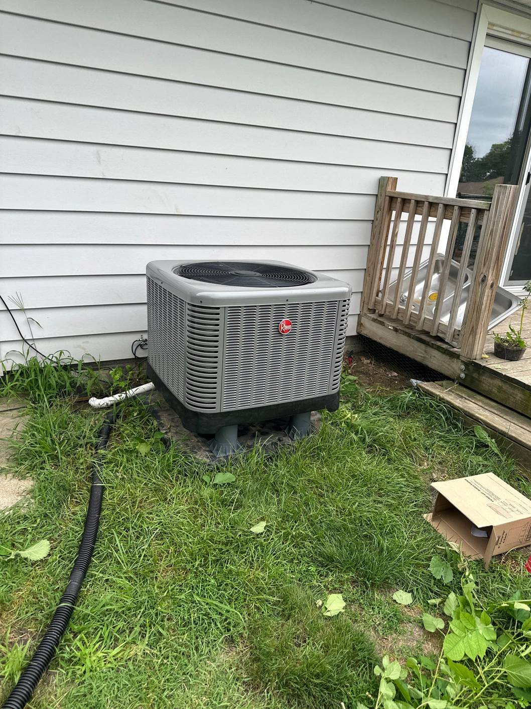 A gray outdoor air conditioning unit sits on a concrete pad next to a white-sided house with a small wooden porch. The surrounding grass is slightly overgrown, and an open cardboard box lies nearby.