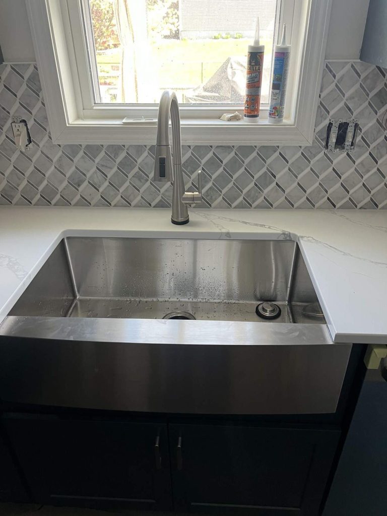 aa36fc7f-e40e-4955-97db-0846b8403428 Stainless steel farmhouse sink with a modern faucet, set in a white countertop. Gray geometric tile backsplash and two caulk tubes are visible on the windowsill above the sink.