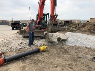 A construction worker guides an excavator lifting broken concrete near a gravel area. A yellow fire hydrant and black pipe lie on the ground in the foreground, with vehicles and buildings in the background.