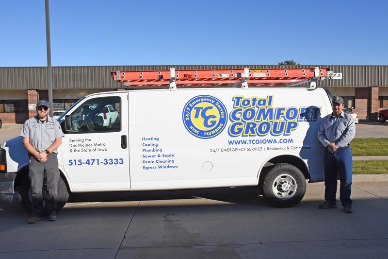 b5ddcab1-57fd-483b-9fca-58665b1386ee Two employees in uniforms stand beside a white Total Comfort Group service van with a red ladder on top, parked outside a commercial building on a sunny day. The van displays company information and services offered.