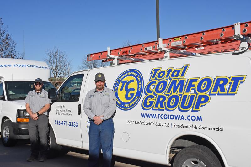 Two uniformed workers stand beside a white van labeled "Total Comfort Group" with blue and yellow branding. The van has a red ladder on top, contact info, and advertises 24/7 emergency service for residential and commercial clients.
