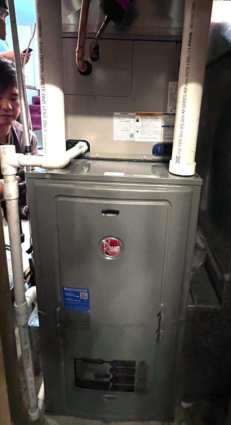 A Rheem furnace with visible PVC pipes attached at the top and side, installed indoors. A person is partially visible on the left side of the image, appearing to inspect the unit.