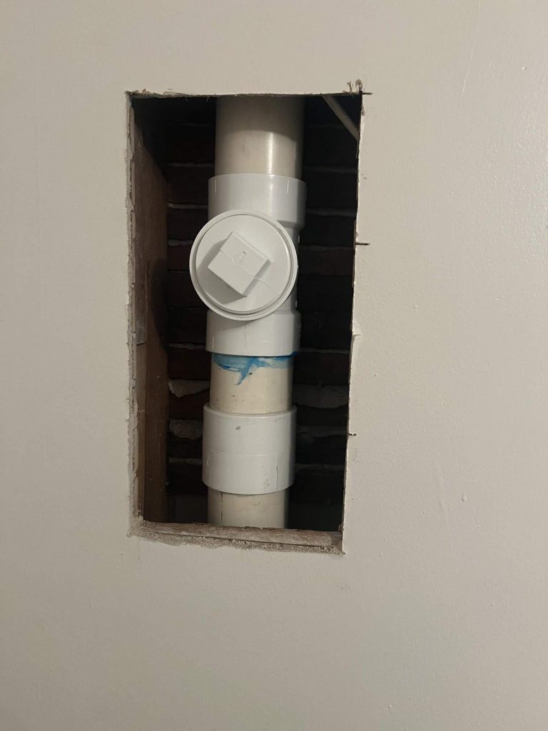 A rectangular hole in a white wall reveals a vertical white PVC pipe with fittings and a cleanout cap, set against a background of exposed brick and wood.