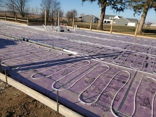 Purple insulation boards cover the ground of a building site, with white radiant heating tubes laid out in loops. Some pipes and utility hookups are visible, and houses and trees can be seen in the background.