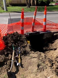 Three orange traffic cones block a hole in the ground next to an orange mesh safety fence. Dirt piles and several shovels are visible, suggesting ongoing construction or repair work. A street and grass are in the background.