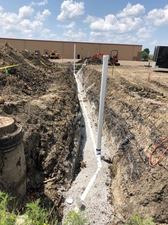 A trench filled with gravel and white PVC pipes runs through a construction site under a partly cloudy sky, with caution tape and machinery visible in the background.