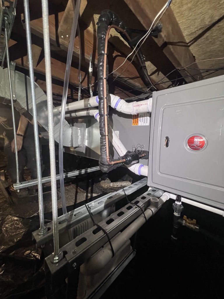 A furnace unit with insulated pipes and wiring installed in an attic space, surrounded by exposed beams and ductwork, with visible support brackets and a Rheem logo on the panel.