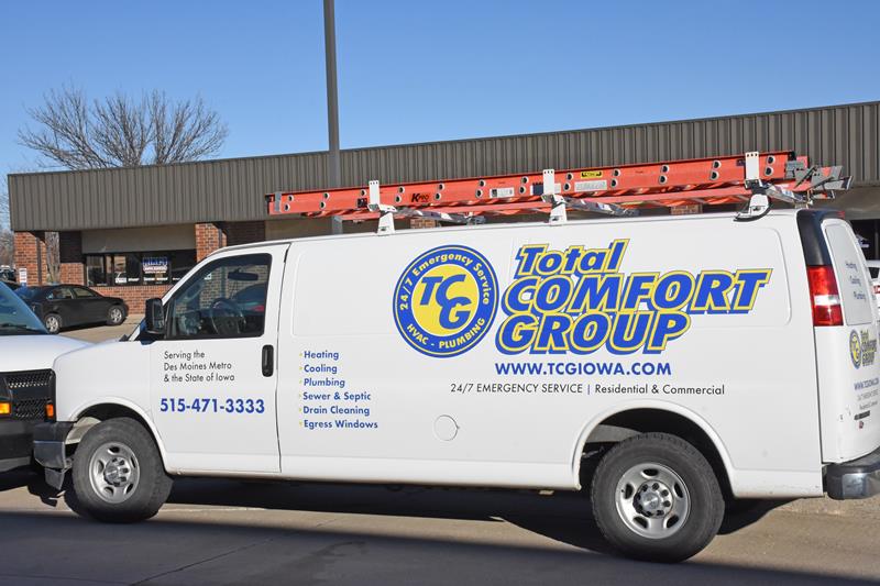 White Total Comfort Group service van with a red ladder on top, parked in front of a building. The van displays company details, services offered, contact number, and website.