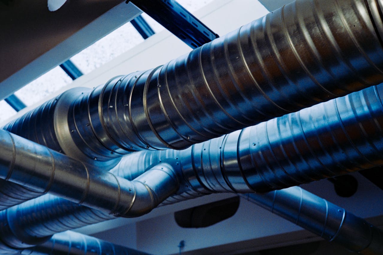 Large silver ventilation ducts run along a ceiling under skylights, with natural light highlighting the metallic surfaces and blue tones. The pipes twist and intersect above an industrial-style interior.